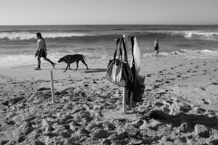 Photo of handbags suspended from a post embedded in a beach, with people walking by in the background