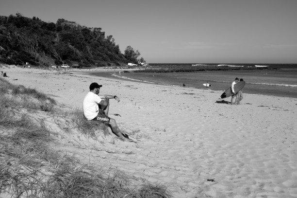 Photo of man sitting on a sandy outcrop watching people on a beach