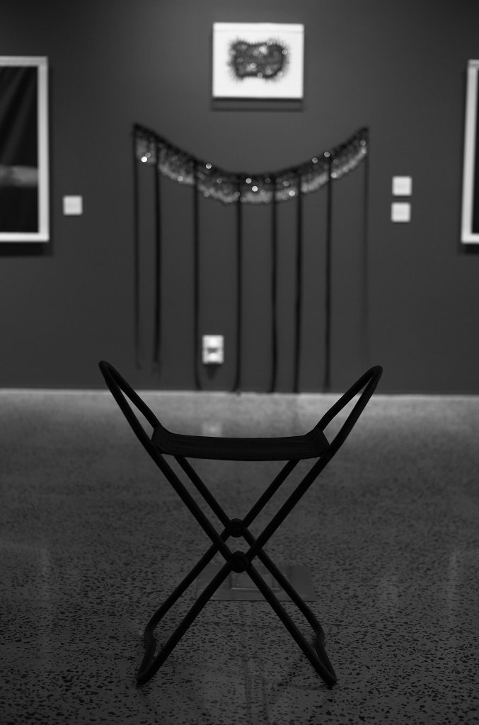 Image of a chair in a dimly lit 'lesser' gallery with an art work on the wall behind the chair.